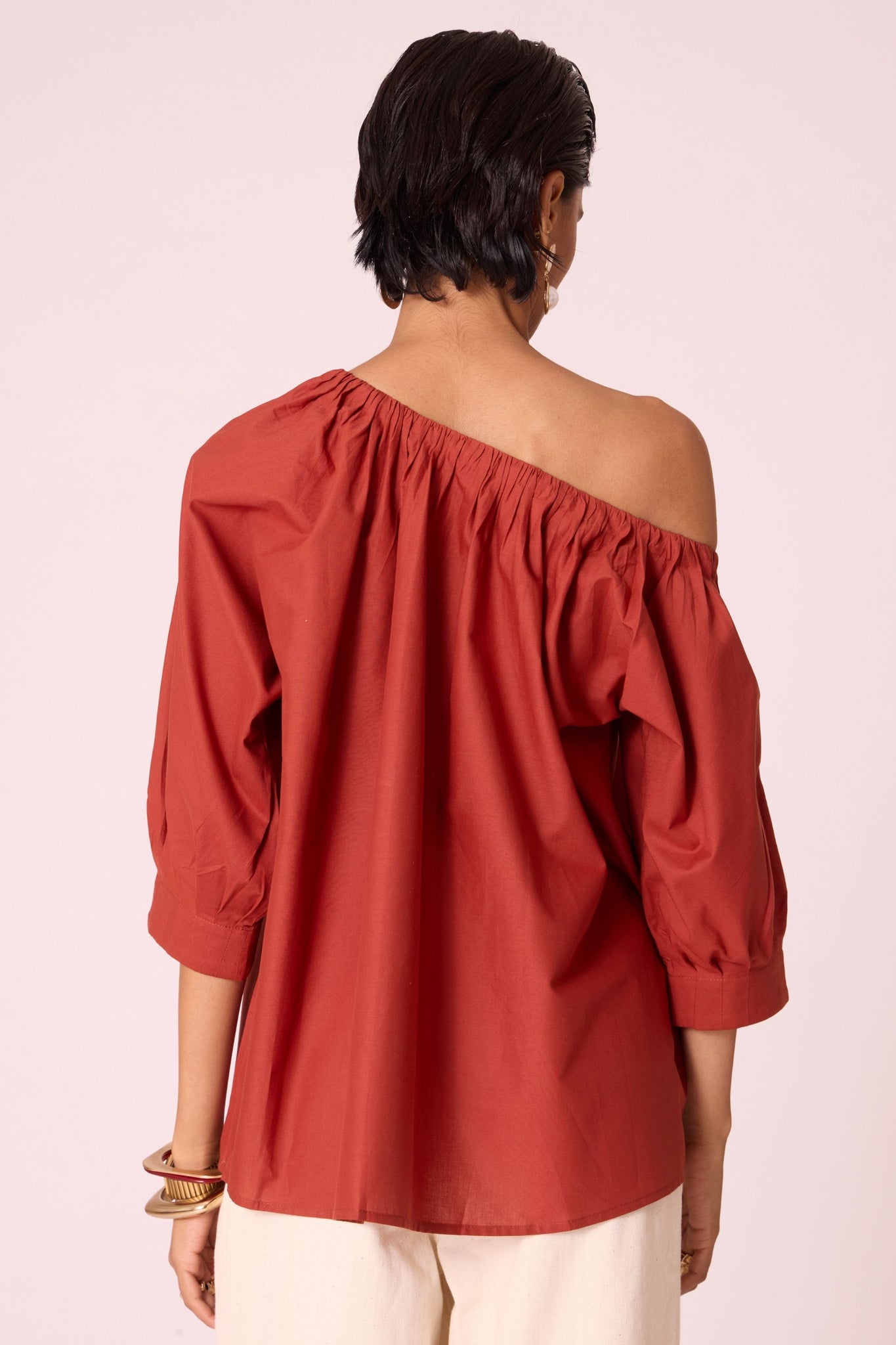 Rust organic cotton off-shoulder top with gathered neckline and pleated sleeve cuffs.