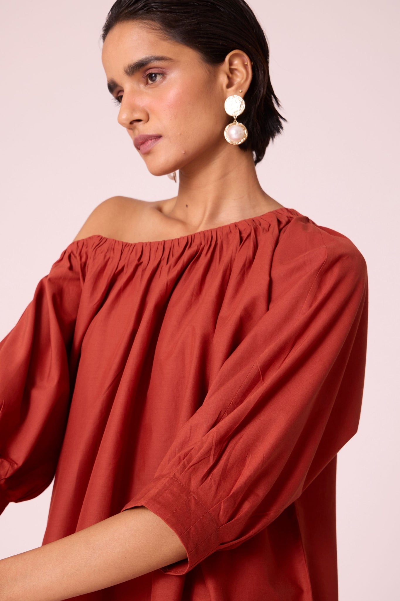 Rust organic cotton off-shoulder top with gathered neckline and pleated sleeve cuffs.
